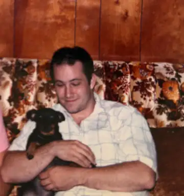 Tony, probably seventeen or eighteen, seated indoors and holding Mindy, a small black-and-tan miniature Doberman.